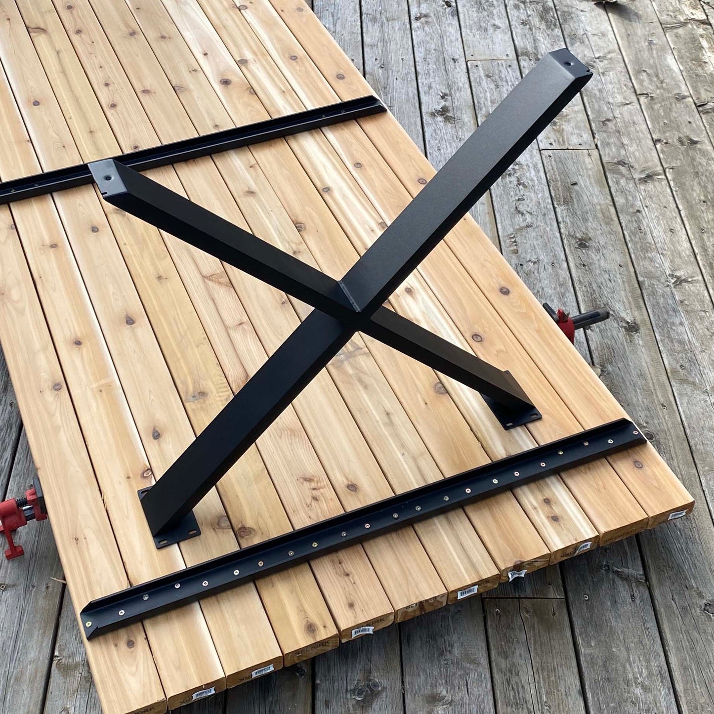A pair of black aluminum X-shaped outdoor dining table legs placed on a wooden surface with visible mounting plates.